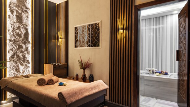 A spa room with a massage table, a stone wall, wooden panels, a window with a view, and a door leading to a bathroom.