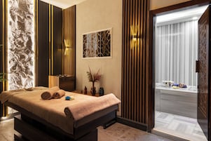 Uma sala de spa com mesa de massagem, parede de pedra, painéis de madeira, janela com vista e porta para um banheiro.
