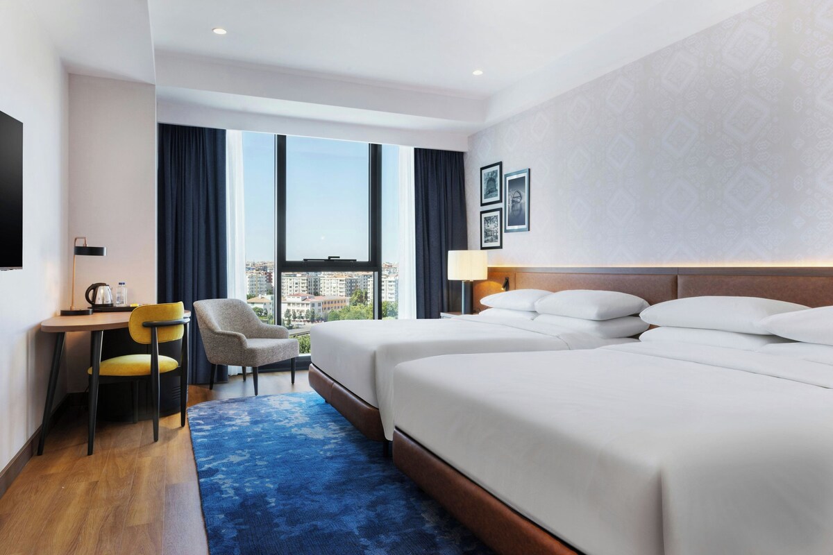 Habitación de hotel con dos camas, un escritorio, una silla y vistas a la ciudad.
