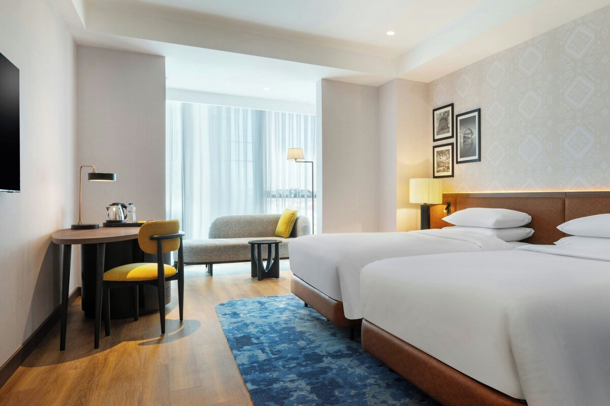 Habitación de hotel con cama, escritorio, sillón, sofá y un amplio ventanal con cortinas.