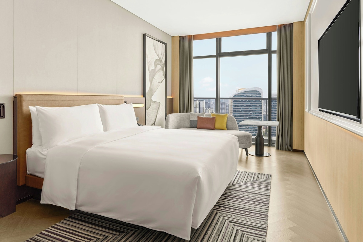 A hotel room with a large bed, a seating area with a table, and a view of the cityscape through large windows.
