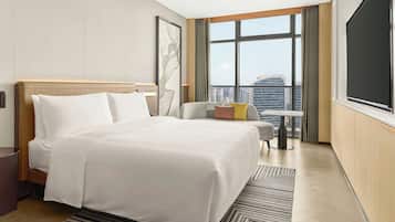 Una camera d'albergo con un letto grande, un'area salotto con un tavolo e vista sulla città attraverso ampie finestre.