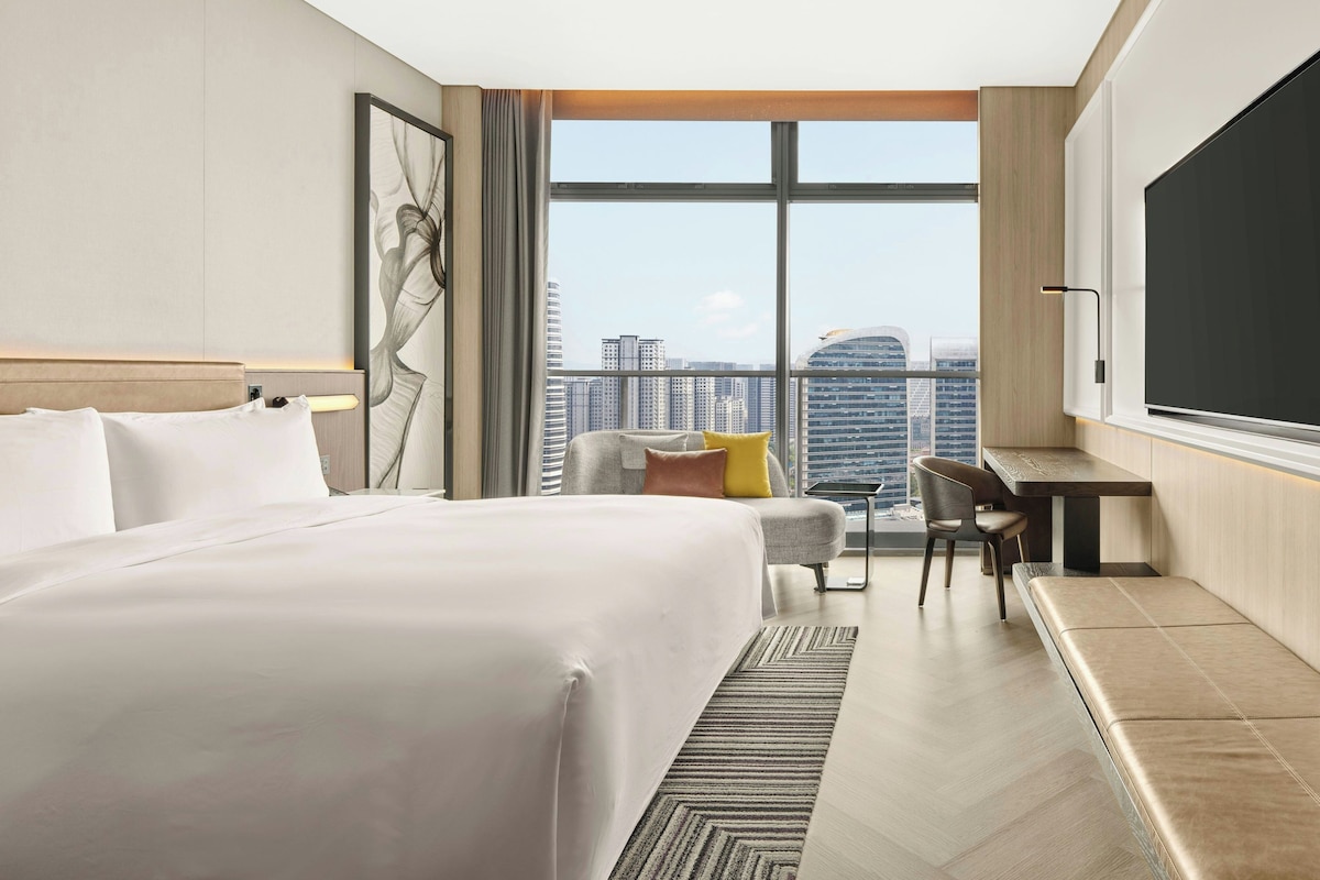 A hotel room with a large bed, a desk, a chair, and a view of the cityscape through large windows.