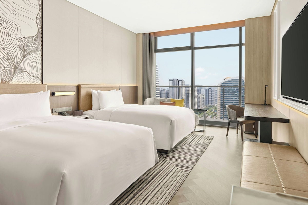A hotel room with two beds, a desk, a chair, and a large window with a city view.