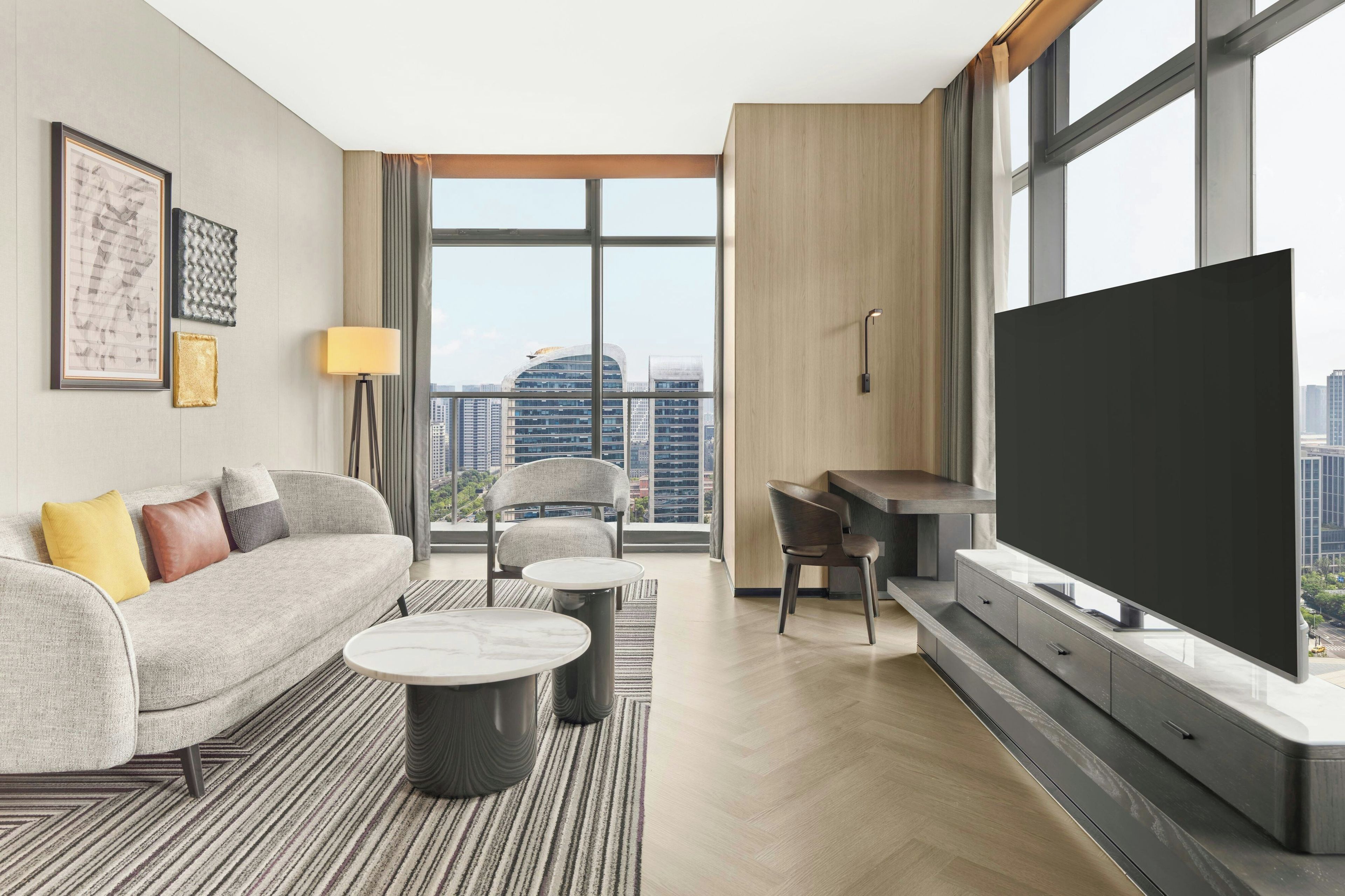 A modern living room with a sofa, coffee table, and chairs, a flat-screen TV, and a city view.