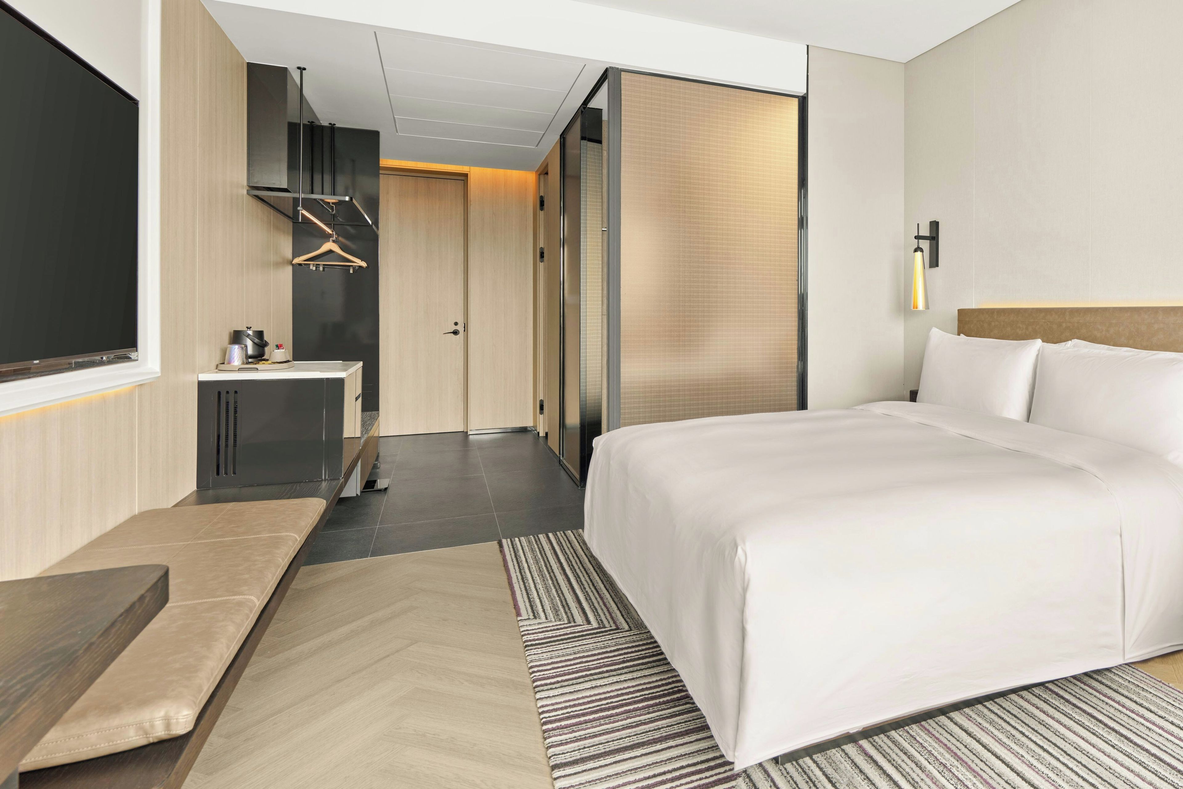 A modern hotel room with a large bed, a bedside table, a desk, and a wardrobe.