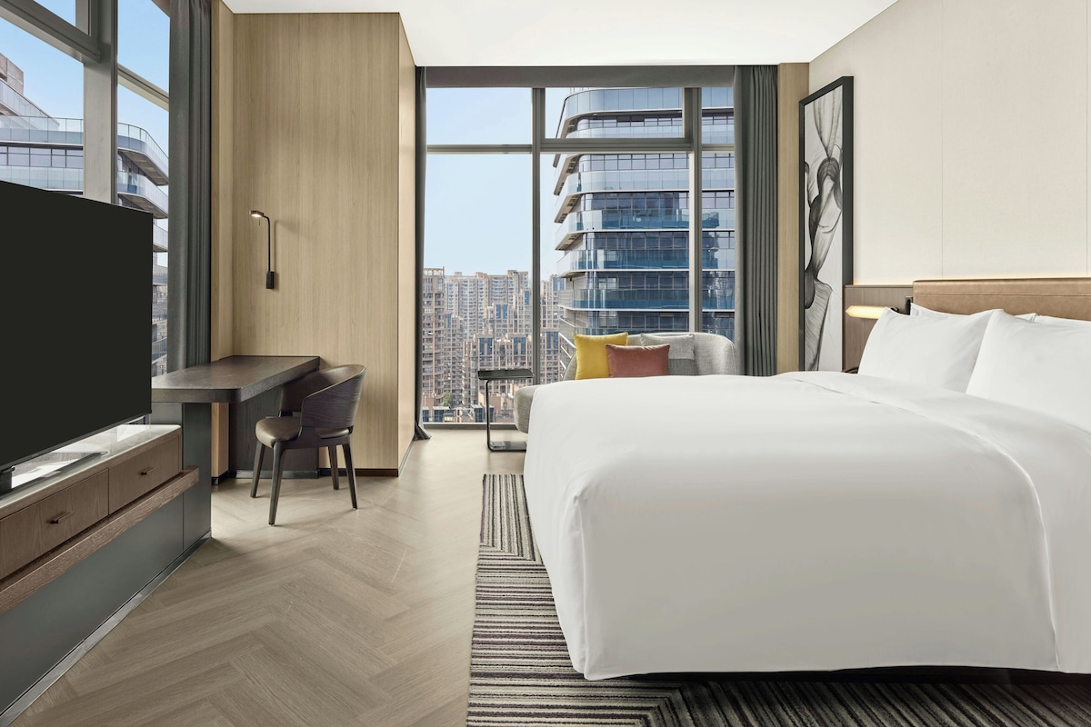 A modern hotel room with a large bed, a desk, a chair, and a view of the cityscape through large windows.