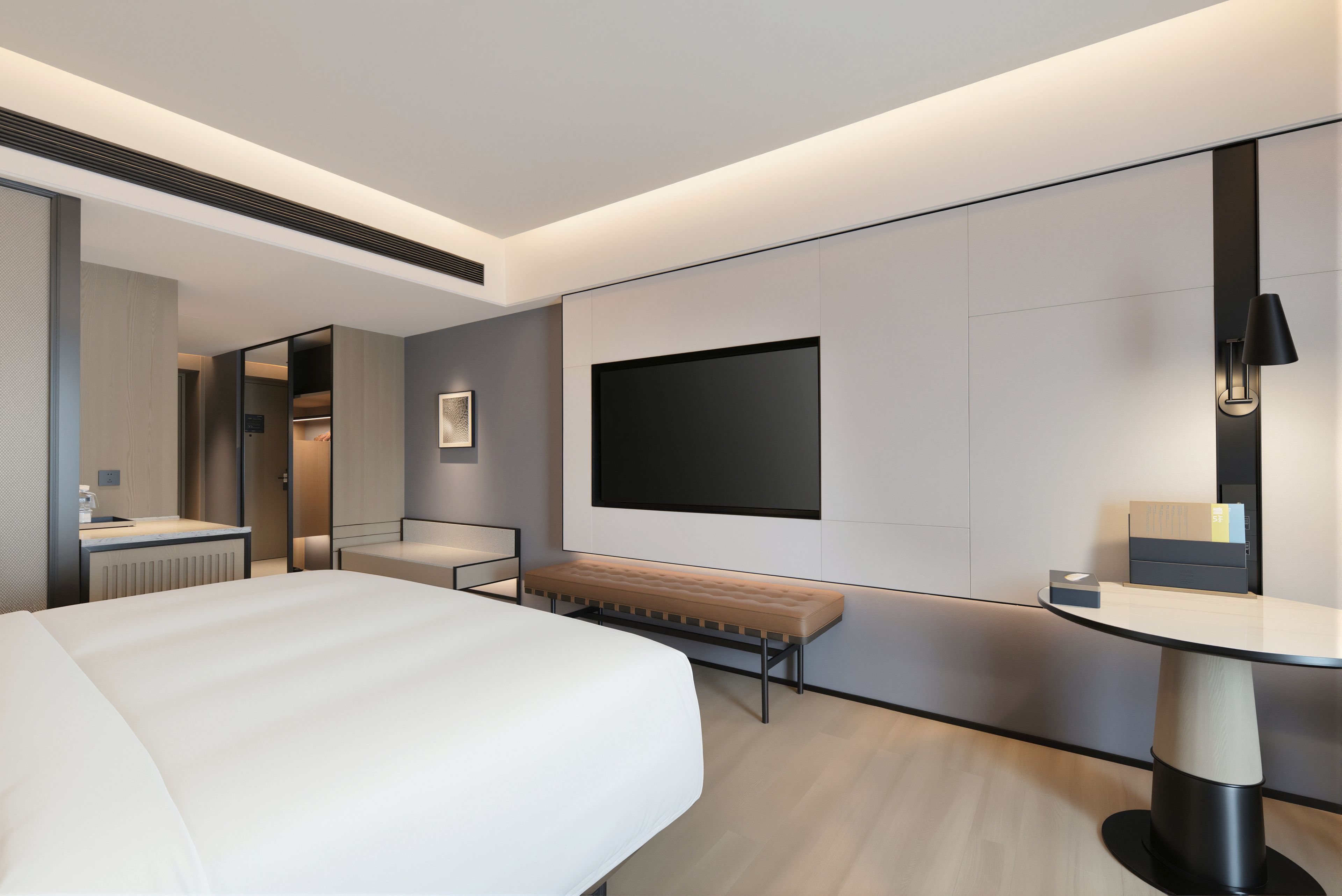 A modern hotel room with a large bed, a bench, a desk, and a flat-screen TV mounted on the wall.
