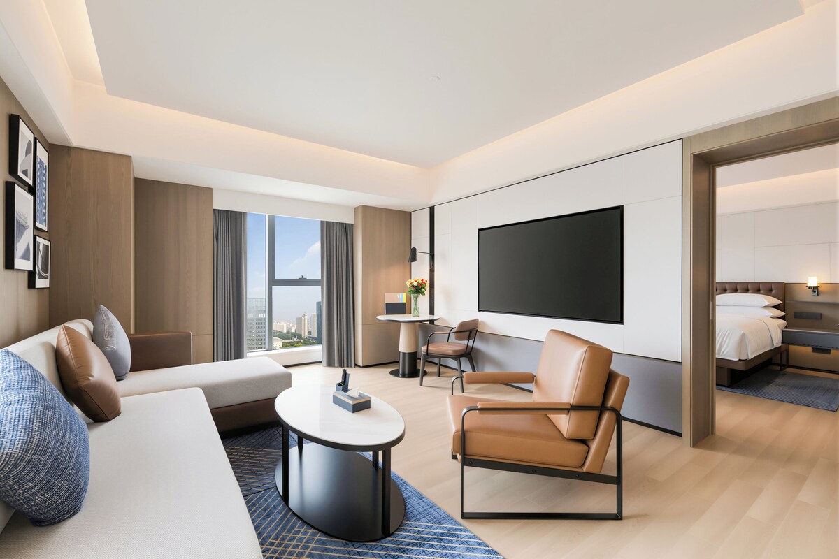 Habitación de hotel moderna con sofá, sillón, televisor de pantalla plana y vista a la ciudad.