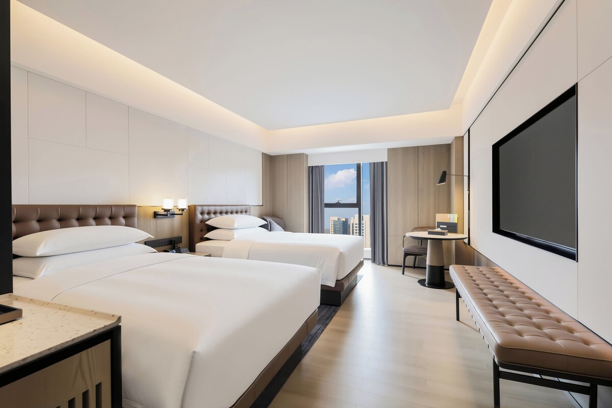 Habitación de hotel con dos camas, un escritorio, un banco, un televisor y vista a la ciudad.