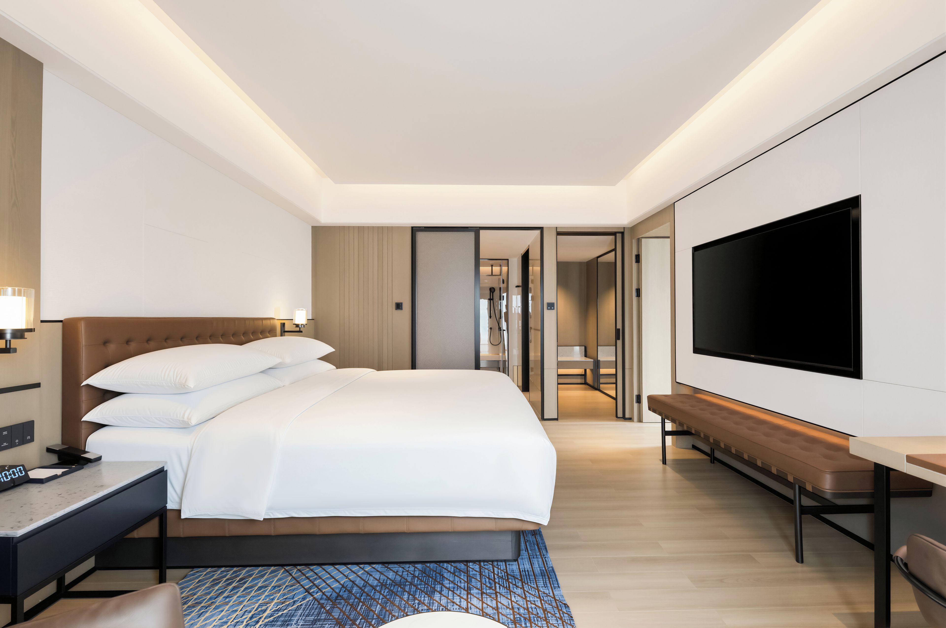 A modern hotel room with a large bed, a flat-screen TV, a bedside table, and a bench.
