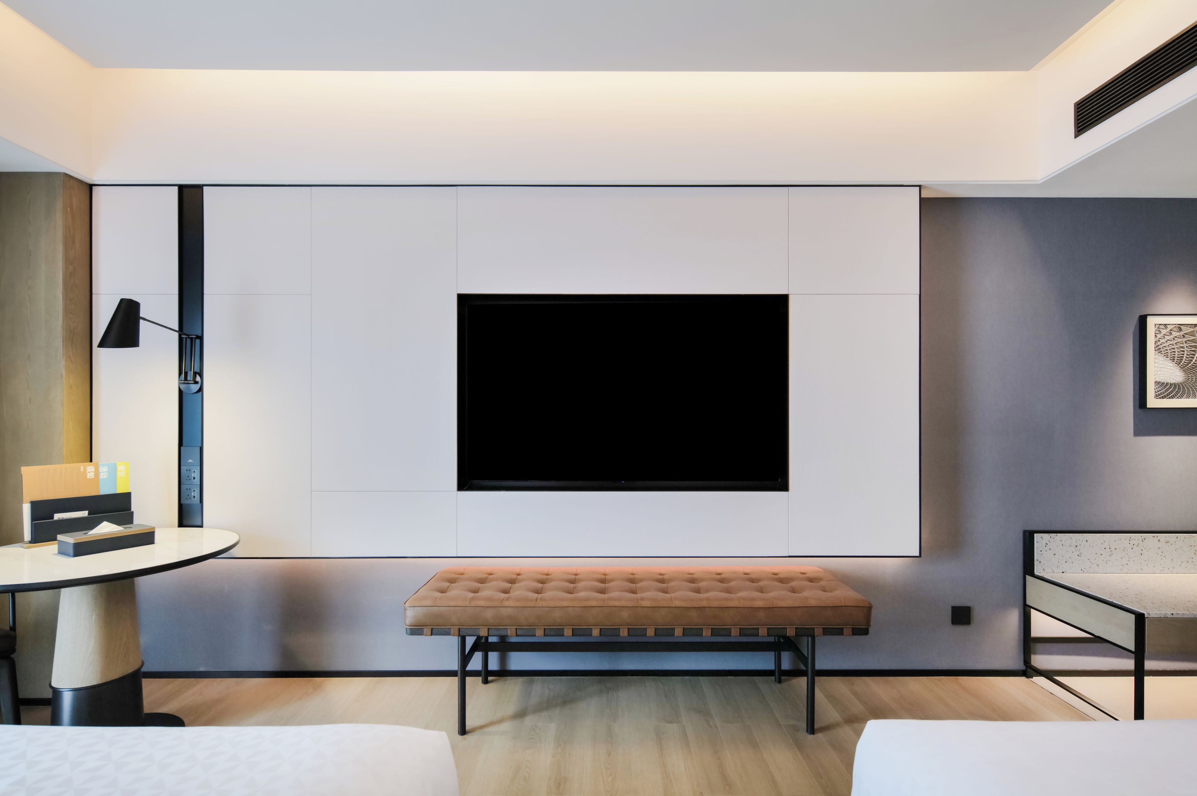 A modern room with a large flat-screen TV mounted on the wall, a round table with a lamp, a bench, and a small sofa.