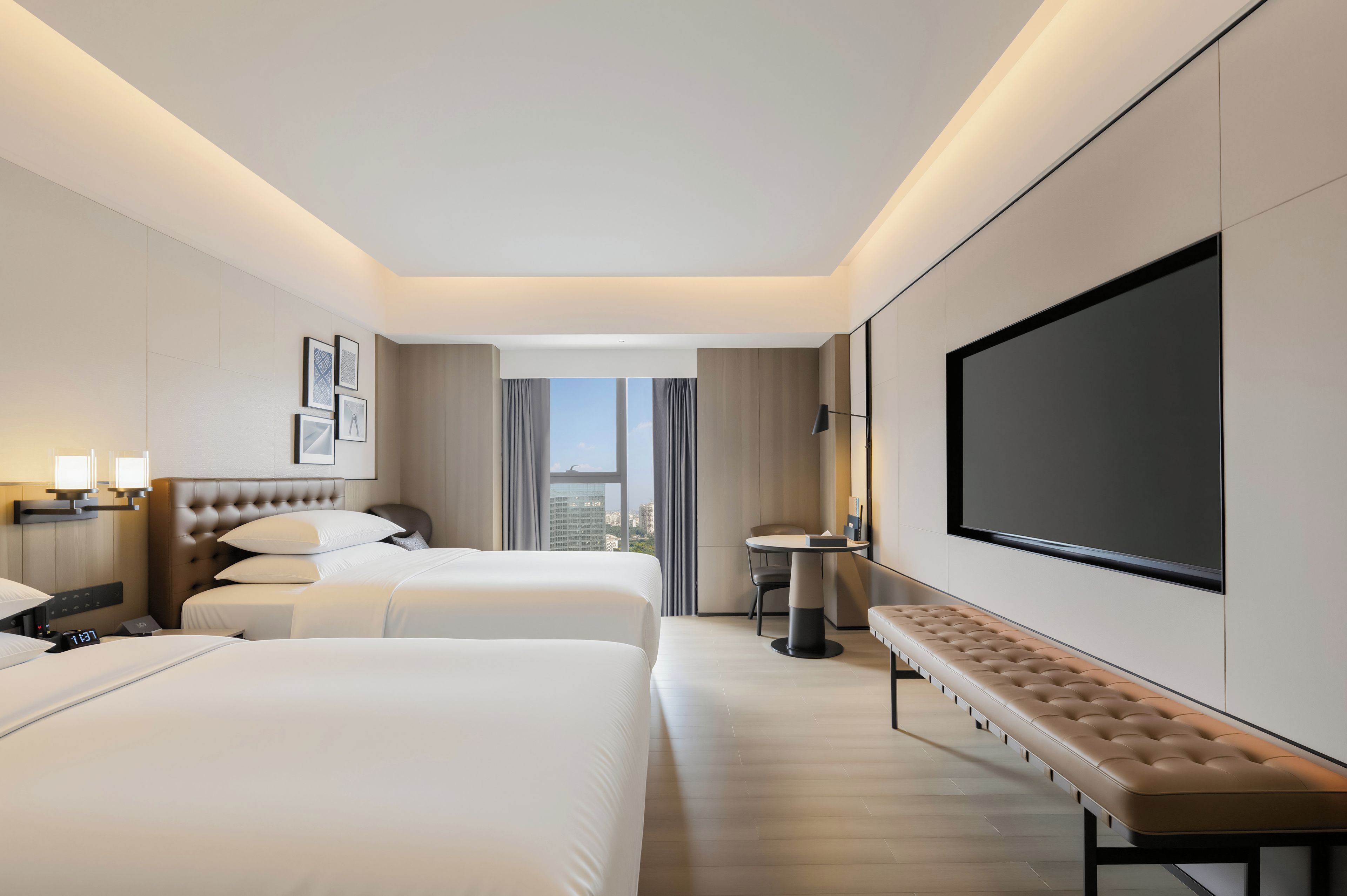A hotel room with two beds, a large TV, a bench, and a view of the city.