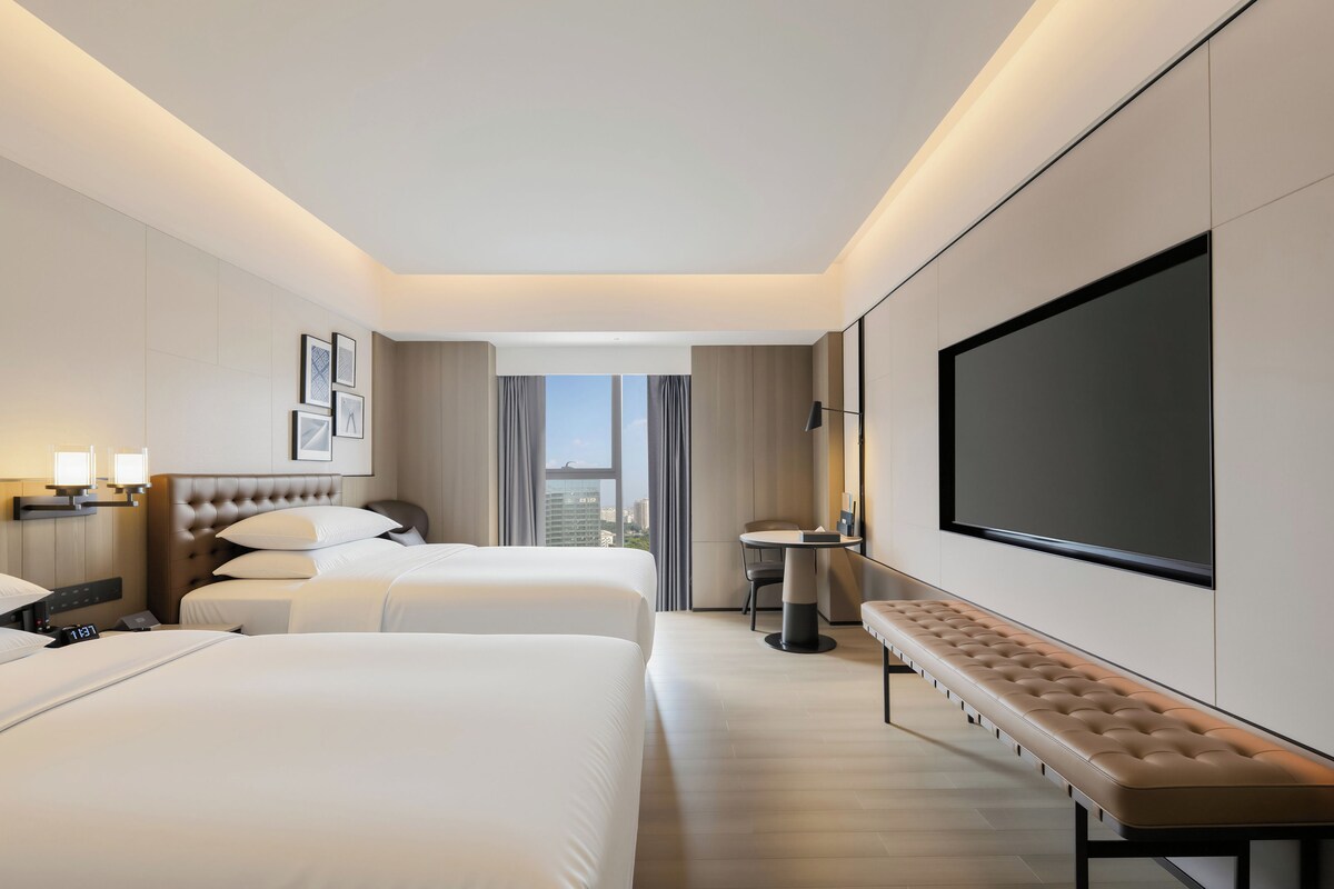Habitación de hotel con dos camas, un televisor grande, un banco y vistas a la ciudad.