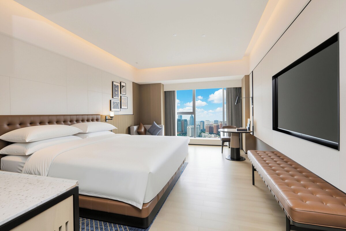 Habitación de hotel con una cama grande, zona de estar, televisor y vista a la ciudad.