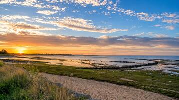 A coastal landscape with a sandy path leading to a body of water, surrounded by grassy areas and a clear sky.