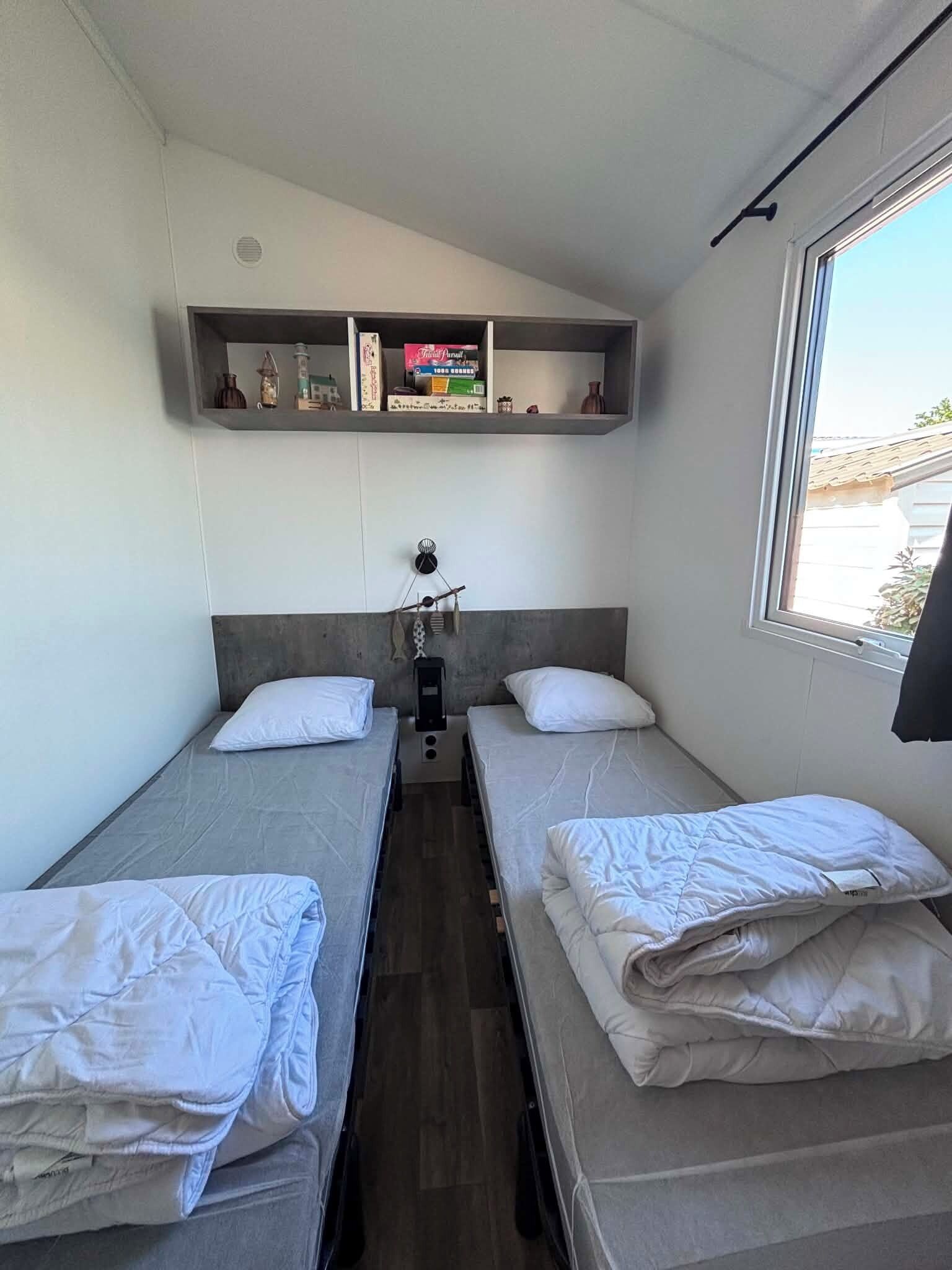 Un petit logement composé d’une seule pièce, comprenant deux lits, une étagère pour les effets personnels, une fenêtre et un radiateur.