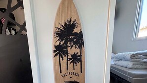 Decoració mural amb forma de taula de surf, amb palmeres i text, situada en una habitació amb llit i finestra.
