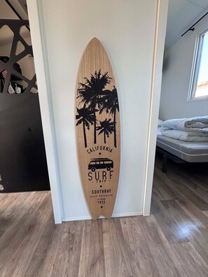 A surfboard-shaped wall decoration with palm trees and text, located in a room with a bed and a window.