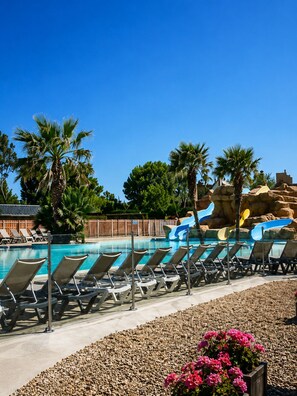 A poolside area with lounge chairs, a water slide, palm trees, and a clear blue sky.