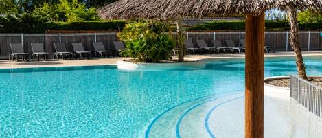 A clear swimming pool with a thatched roof structure, surrounded by lounge chairs and a fence.