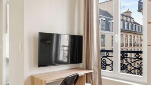 A room with a flat-screen TV mounted on a wall, a wooden desk, and a window with a view of a cityscape. - Appartement de charme rue des Mauvais Garcons (Paris)