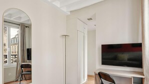 A modern living room with a flat-screen TV, a dining area with a chair, and a view of a building through an archway. - Appartement de charme rue des Mauvais Garcons (Paris)