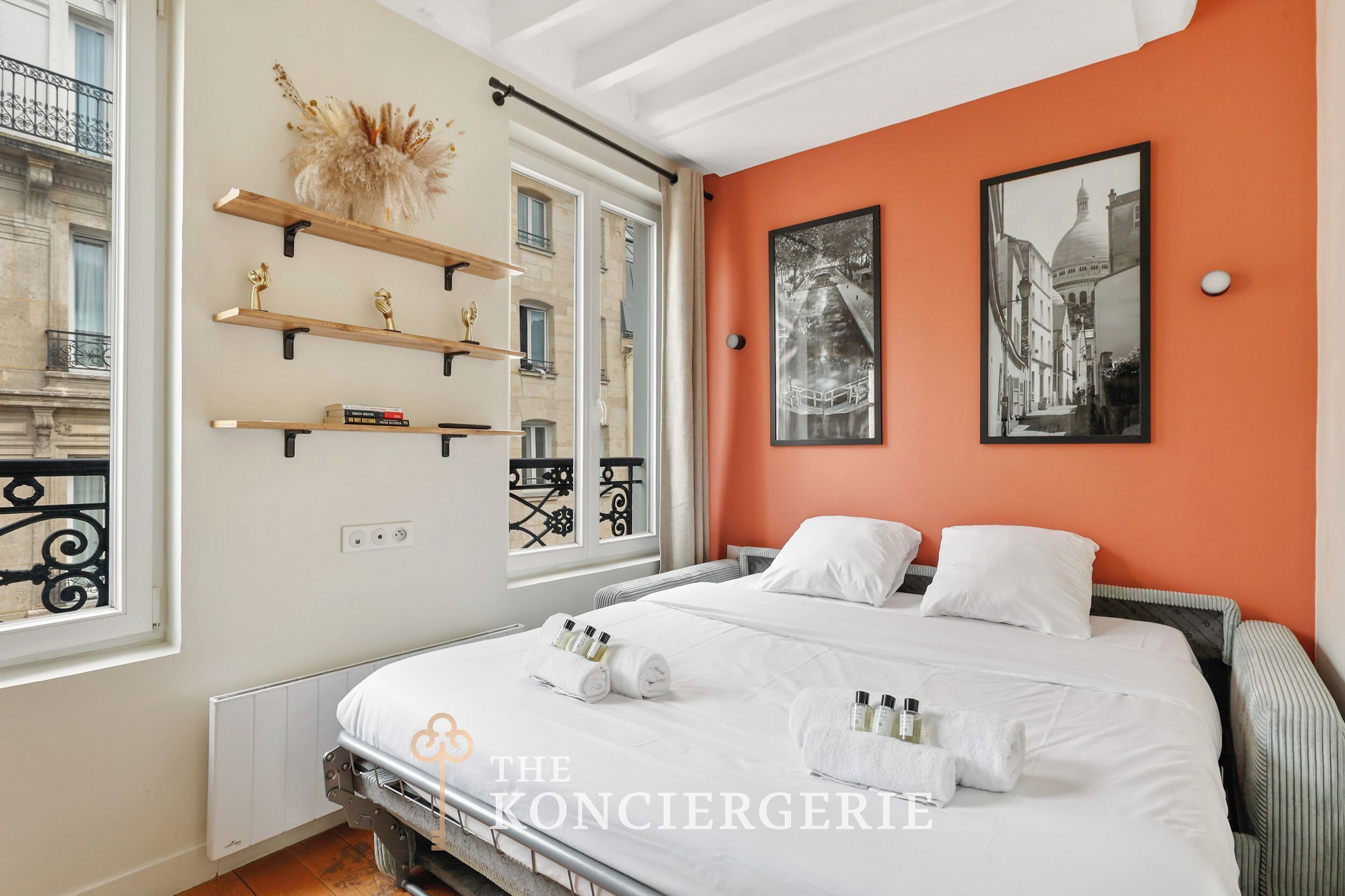 Ein Hotelzimmer mit einem Bett, zwei Kissen, einem Nachttisch, einem Fenster mit Blick auf Gebäude und einer Wand mit zwei gerahmten Bildern.