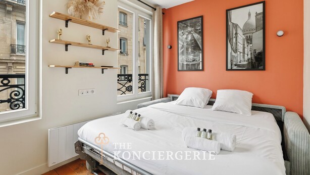 A hotel room with a bed, two pillows, a nightstand, a window with a view of buildings, and a wall with two framed pictures. - Appartement de charme rue des Mauvais Garcons (Paris)