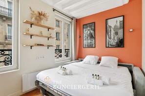A hotel room with a bed, two pillows, a nightstand, a window with a view of buildings, and a wall with two framed pictures. - Appartement de charme rue des Mauvais Garcons (Paris)