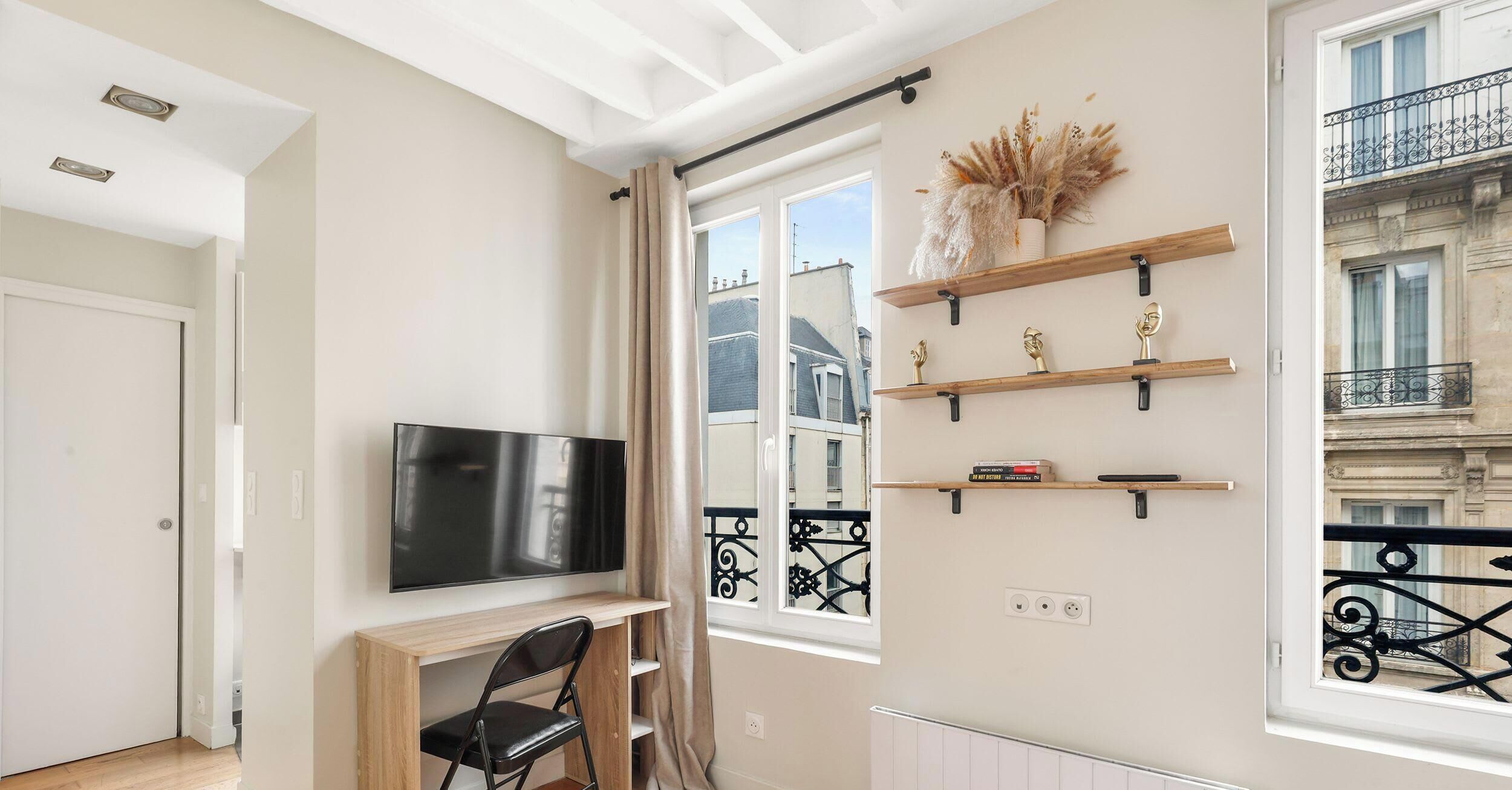 Un salon moderne doté d’un téléviseur à écran plat, de meubles en bois, d’objets décoratifs disposés sur des étagères et offrant une vue sur la ville grâce à de grandes fenêtres.
