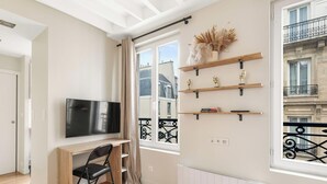 A modern living room with a flat-screen TV, wooden furniture, decorative items on shelves, and a view of the city through large windows. - Appartement de charme rue des Mauvais Garcons (Paris)