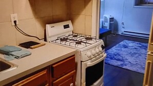 Een keuken met een fornuis, oven en houten keukenkastjes. Er is een raam en een deur die naar een andere kamer leidt.