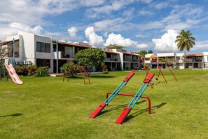 Ein Spielplatz mit Schaukeln und einer Rutsche, umgeben von einem Gebäude mit Balkonen und blauem Himmel.