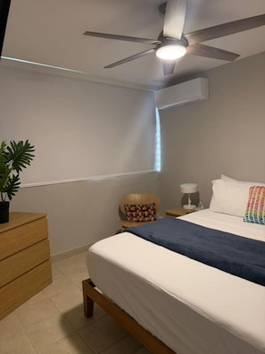 Ein Schlafzimmer mit einem Bett, einem Deckenventilator, einem Nachttisch, einer Lampe, einem Sessel und einer Topfpflanze.