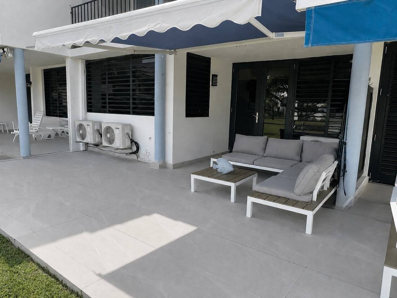 A covered outdoor seating area with a sofa and a table, air conditioning units, and a building with a blue awning.