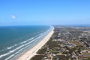 An aerial view of a coastal town with a long beach, residential areas, and a clear view of the ocean.