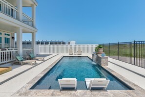 A rectangular swimming pool with a white deck, two lounge chairs, a white fence, and a view of a building in the background. - Blue Haven Retreat – Coastal Luxury Steps to Beach (Port Aransas)