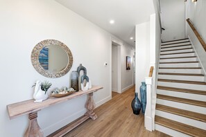 A hallway with a wooden console table, decorative vases, and a mirror with a beach scene. A staircase with wooden steps and a white railing is visible to the right. - Luxurious Retreat. Ocean Views. Steps to the Beach (Port Aransas)
