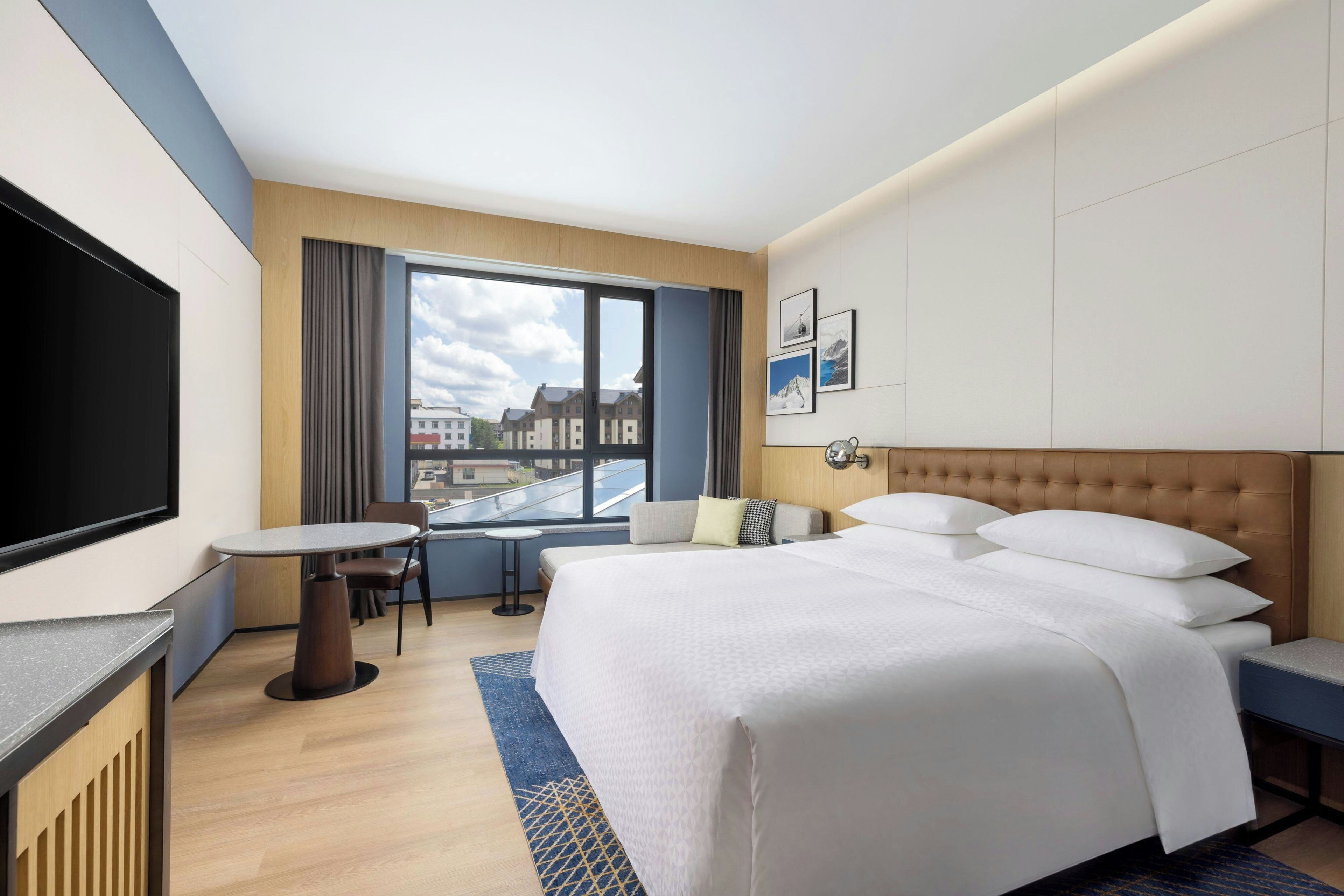 Quarto de hotel com cama, televisão, escrivaninha e janela com vista para edifícios.