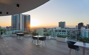 En tagterrasse med udsigt over byen i solnedgang, med et bord og stole, plantede planter og moderne bygninger.