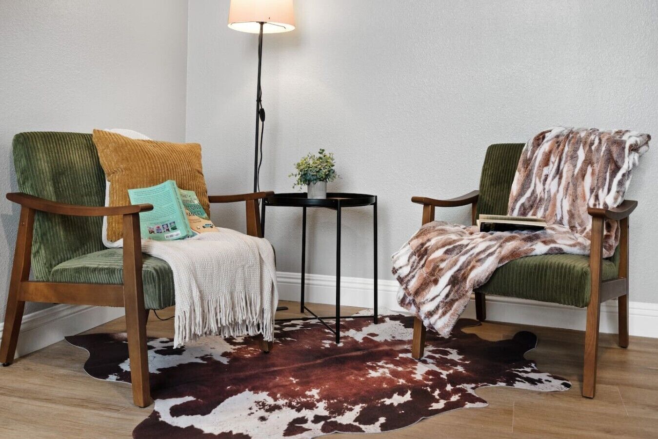Une pièce comprenant deux fauteuils verts, une petite table, un tapis en peau de vache et un lampadaire.