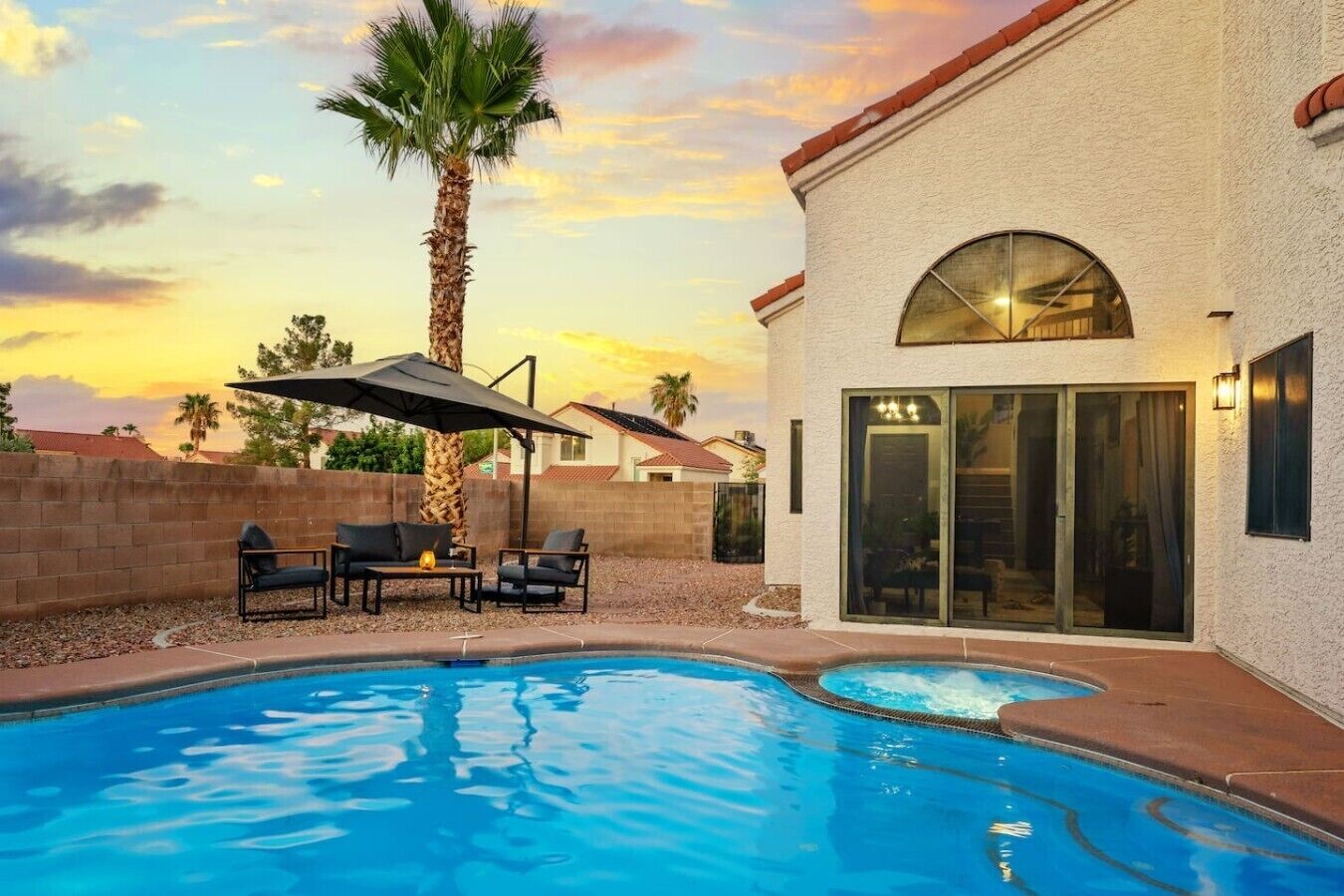 Piscina con área de patio, que incluye sillas y un paraguas, junto a un edificio con un gran ventanal y una puerta.