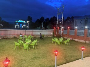 An outdoor seating area with green chairs and tables, a fenced perimeter, and a building with illuminated signage in the background. - Hotel Classy Castle (Pahalgam)