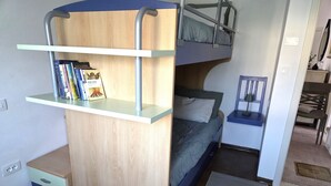 Une petite pièce avec un lit superposé, un bureau avec des livres, un petit tiroir et un tapis sur le sol.