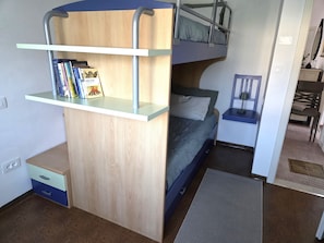 Ein kleiner Raum mit Etagenbett, Schreibtisch, Büchern, einer kleinen Schublade und einer Matte auf dem Boden.