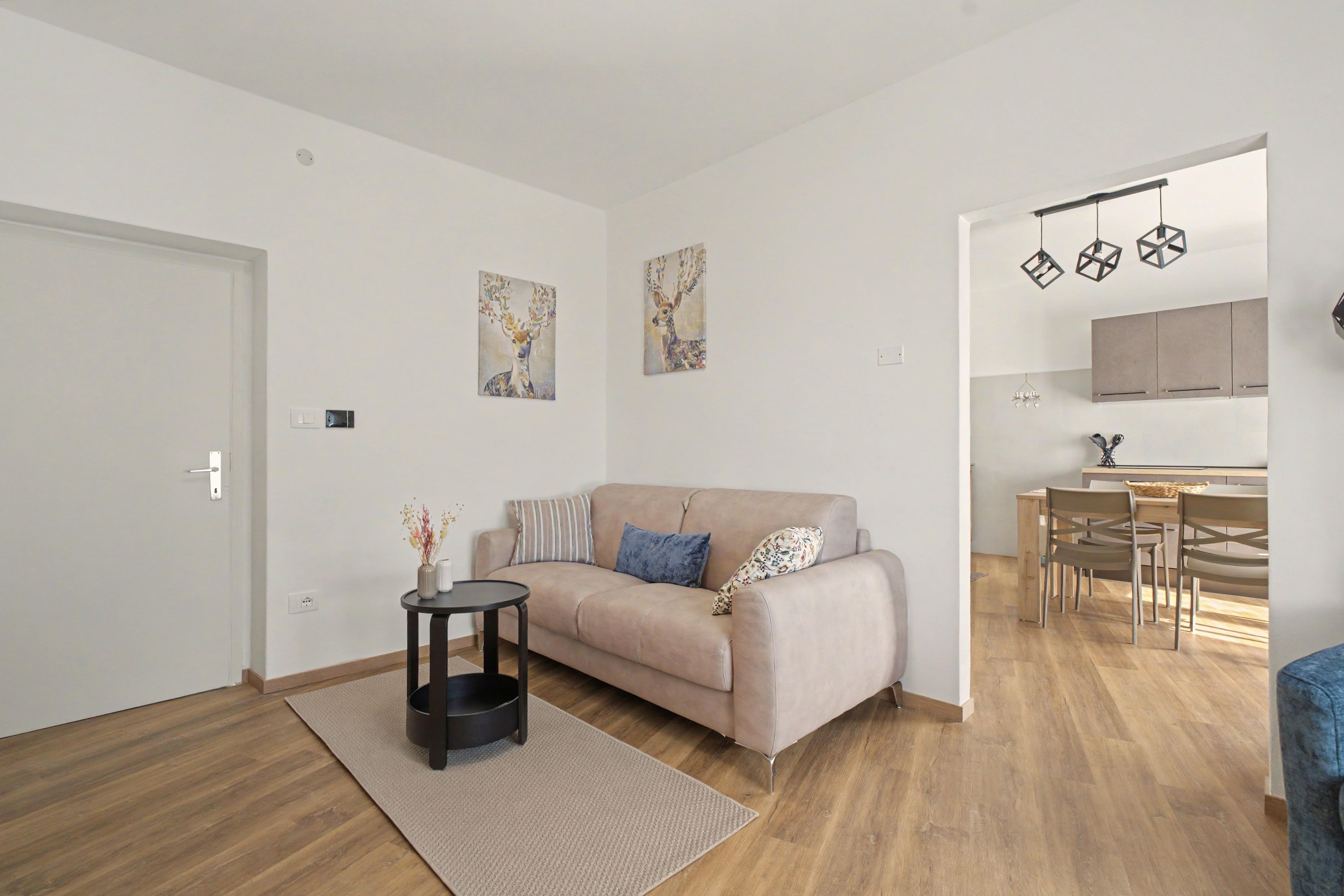 Ett modernt vardagsrum med en beige soffa, ett litet sidobord och en matta. I anslutning till vardagsrummet finns ett köksområde med ett matbord och stolar, som syns genom en öppen dörröppning.