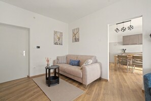 Ein modernes Wohnzimmer mit einem beigen Sofa, einem kleinen Beistelltisch und einem Teppich. Durch einen offenen Durchgang ist eine Küchenzeile mit Esstisch und Stühlen zu sehen.
