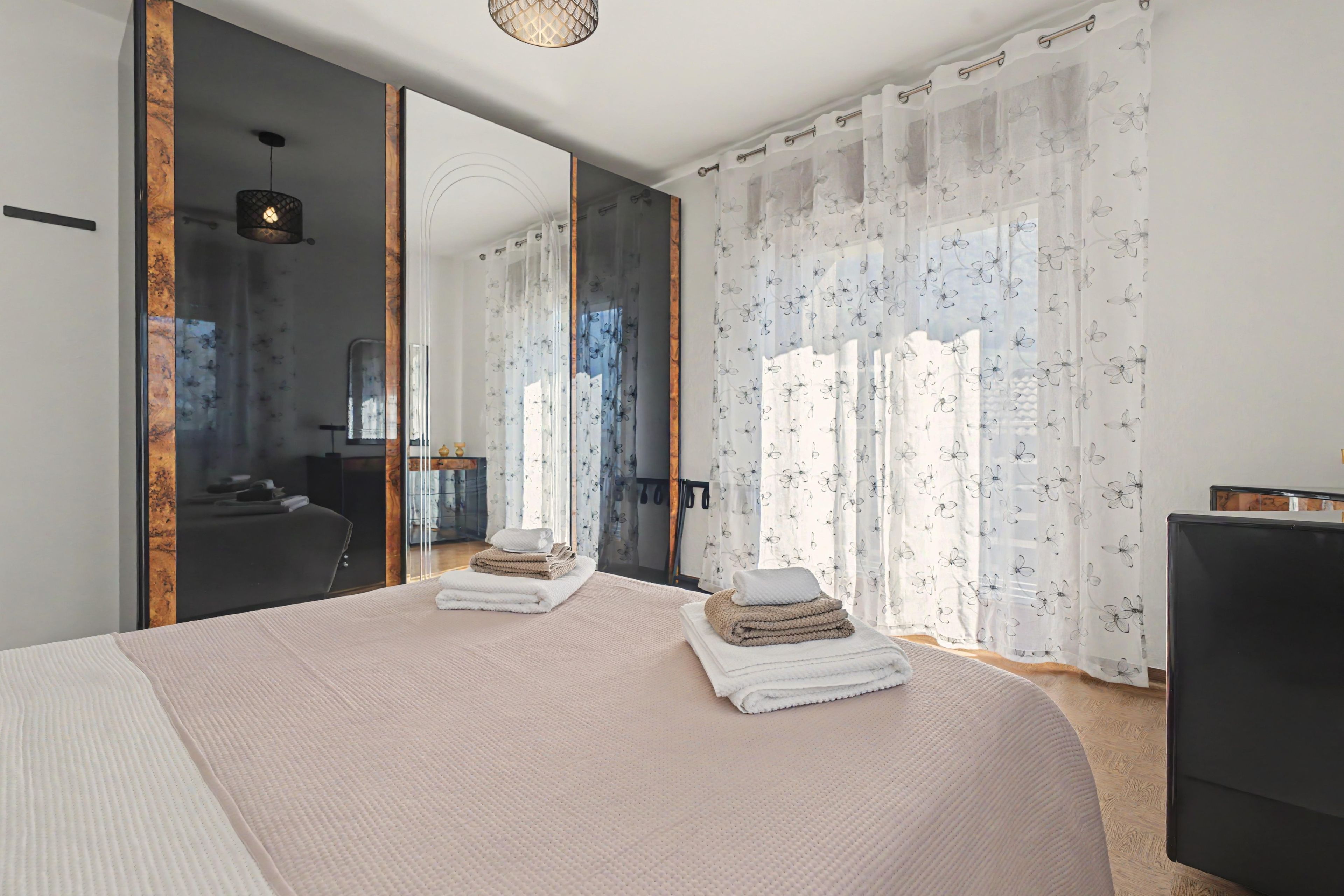 A modern bedroom with a large bed, a mirror, and a view of a cityscape through sheer curtains.