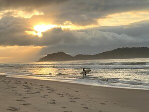 A beach with footprints leading to the water, a person carrying a surfboard, and a sunset over the ocean. - Modern retreat in Novo Campeche, 100m from the beach! (Florianópolis)